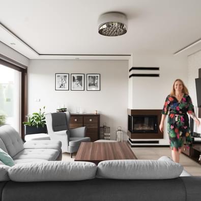 Jasny salon z szarym narożnikiem, drewnianym stolikiem kawowym, kominkiem z czarnymi akcentami i kobietą w kwiecistej sukience w tle.