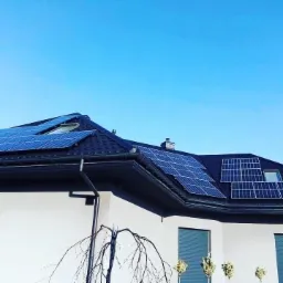 Dom jednorodzinny z ciemnym dachem pokrytym panelami słonecznymi, widoczne okno dachowe i komin, na pierwszym planie ozdobne drzewko bez liści.