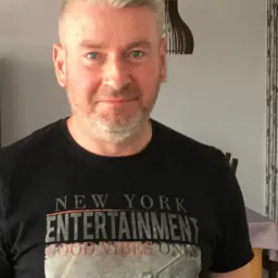 Portret mężczyzny z blond włosami i niebieskimi oczami, ubranego w czarną koszulkę z nadrukiem 'New York Entertainment', na tle jasnej ściany z drewnianą lampą.