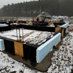 Fundamenty domu jednorodzinnego w trakcie budowy, ocieplone styropianem, z widocznymi zbrojeniami i pracownikami w oddali, zimowy krajobraz.