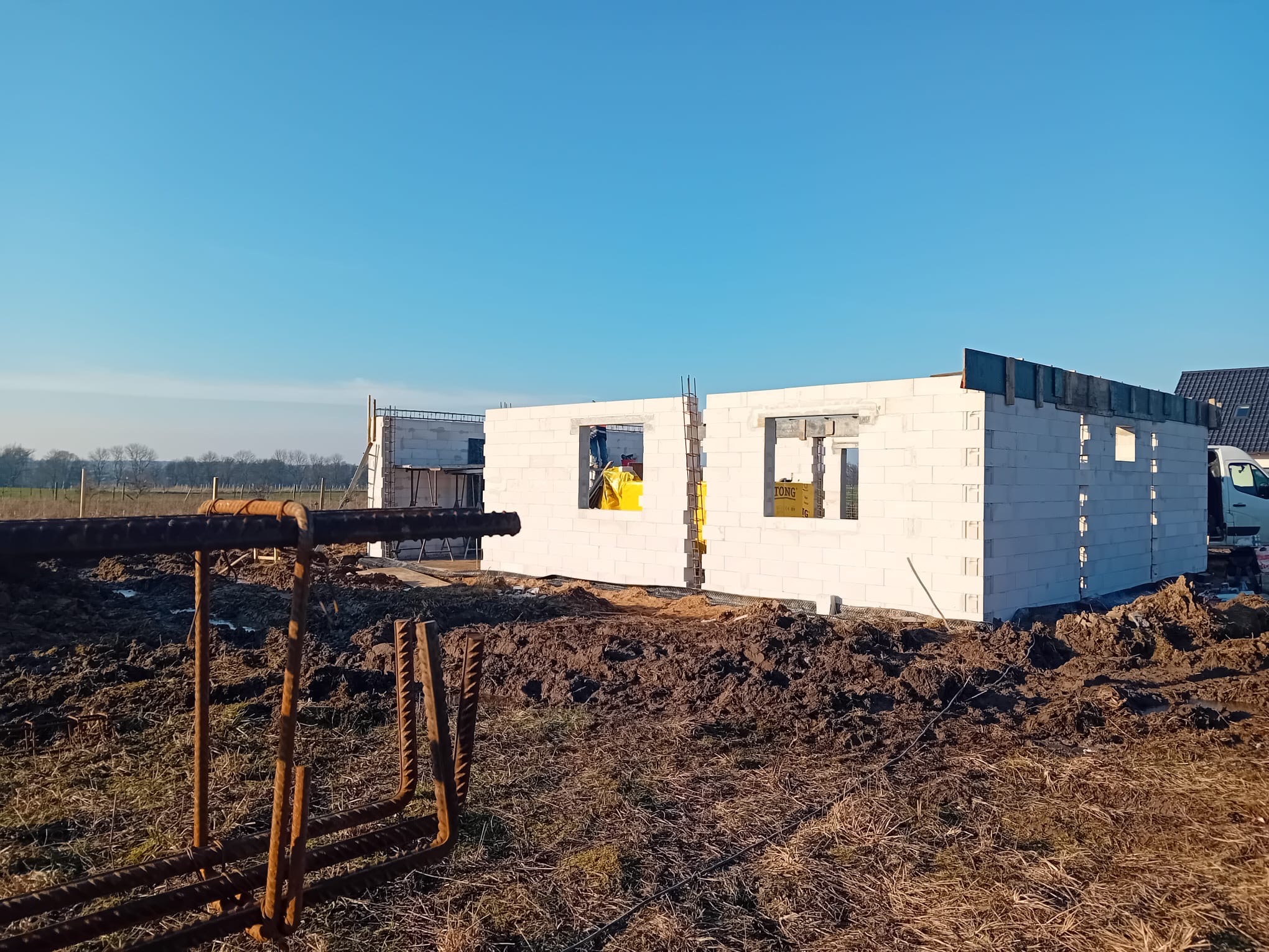 Wznoszenie ścian parterowego domu z bloczków betonu komórkowego; widoczne zbrojenia i żółta folia zabezpieczająca okna.
