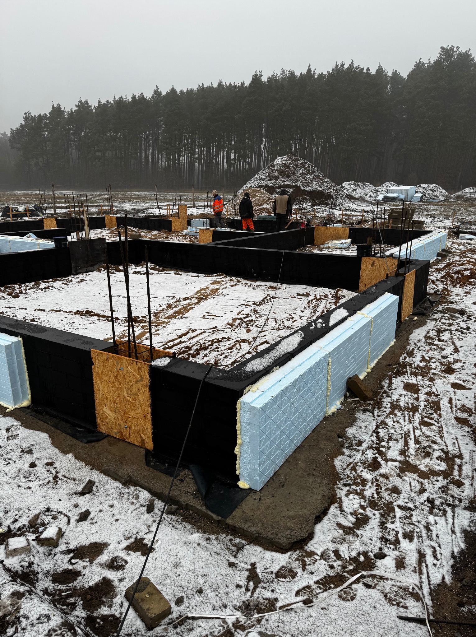 Fundamenty domu jednorodzinnego w trakcie budowy, ocieplone styropianem, z widocznymi zbrojeniami i pracownikami w oddali, zimowy krajobraz.