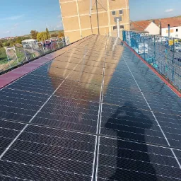 Rząd paneli słonecznych na dachu budynku przemysłowego, zabezpieczony siatką ochronną, z widokiem na okoliczne budynki i turbiny wiatrowe w oddali.