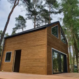 Dwukondygnacyjny budynek z drewnianą elewacją, z czarną ramą okienną i dachem, otoczony wysokimi drzewami i drewnianym tarasem w trakcie budowy.