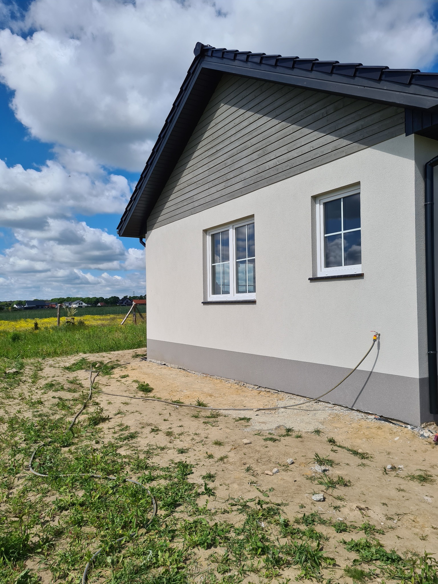 Nowoczesny dom z szarą drewnianą elewacją, jasnym tynkiem i ciemną dachówką, widok z boku na tle kwitnącej łąki i błękitnego nieba z chmurami. Wokół domu widoczny niezagospodarowany teren...