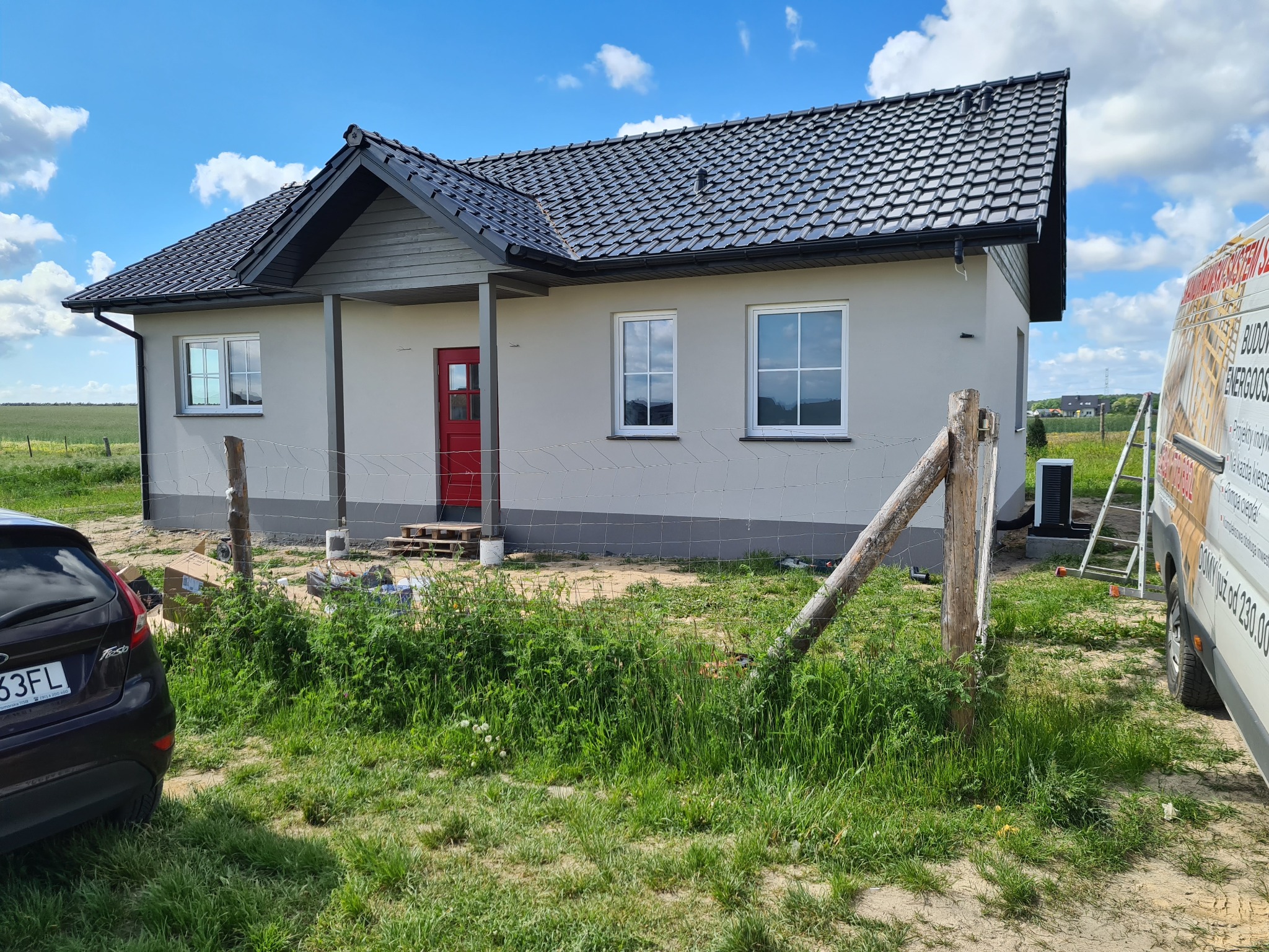 Gotowy, parterowy dom z szarą elewacją, czarną dachówką i czerwoną stolarką drzwiową, widziany z perspektywy niskiego ogrodzenia z drewnianymi słupkami i samochodem osobowym w tle, na tle zielonego...