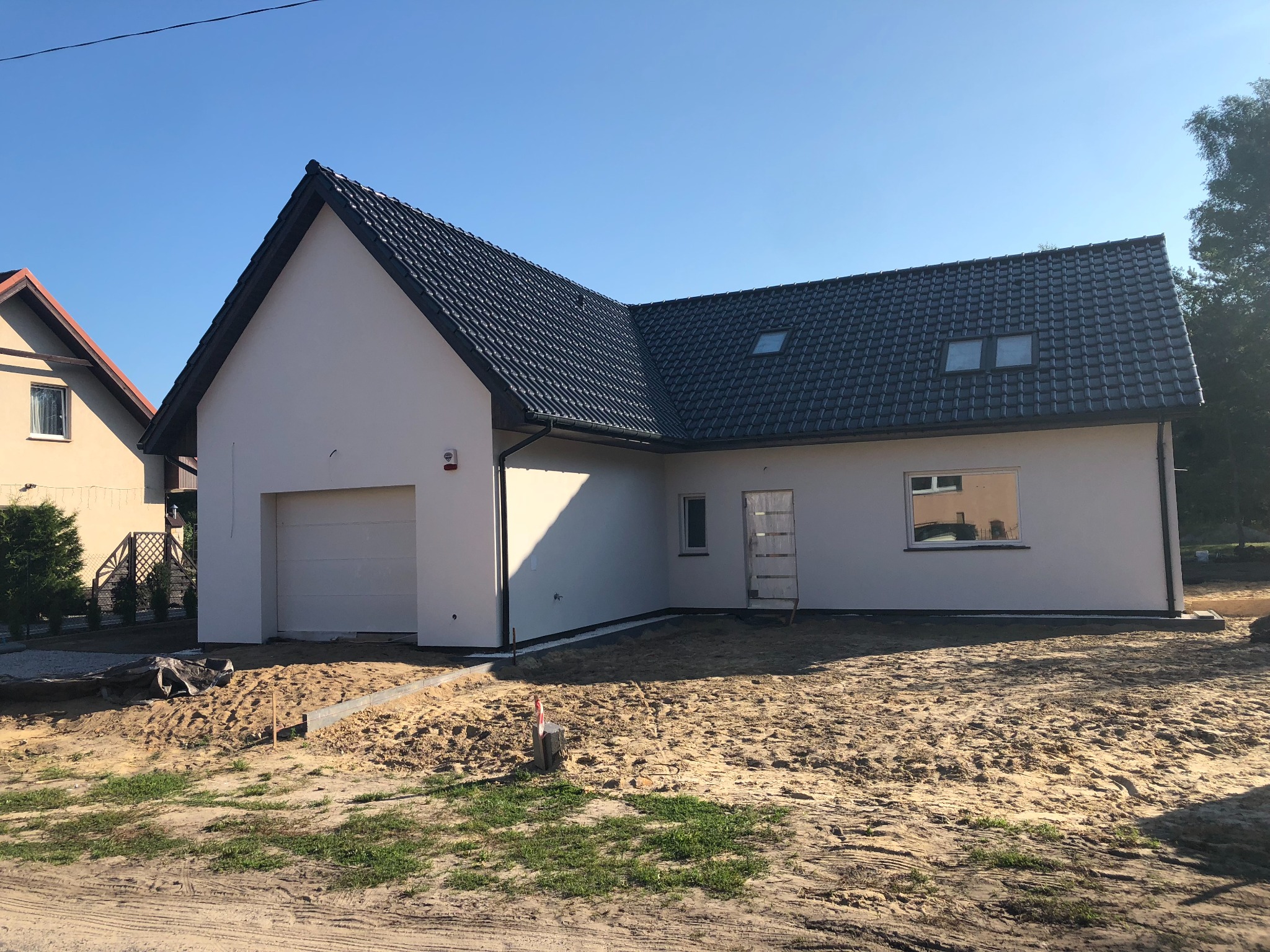 Nowoczesny dom jednorodzinny z garażem w trakcie budowy, widoczne okna dachowe, czarna dachówka i jasna elewacja, teren wokół domu przygotowany pod zagospodarowanie.