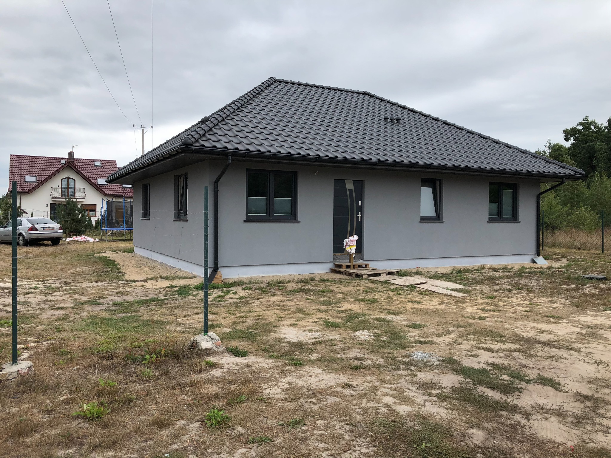 Nowoczesny dom parterowy z szarą elewacją i ciemną dachówką karpiówką, widoczny z zewnątrz na działce w trakcie zagospodarowywania, z tymczasowym wejściem z palet.