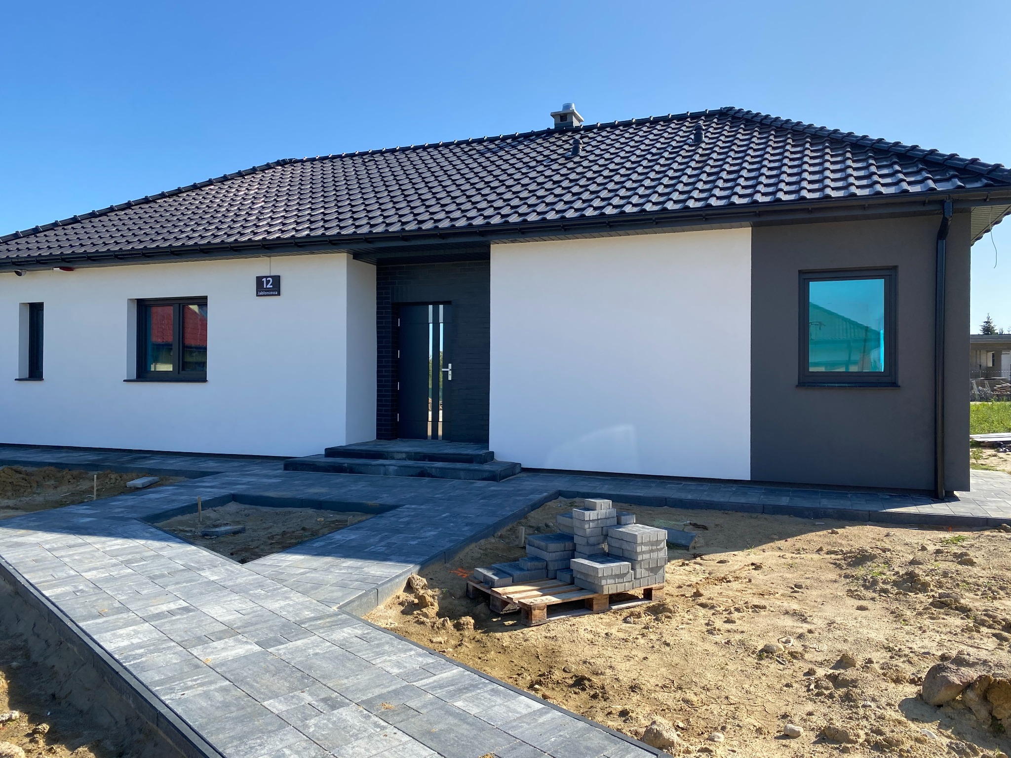 Nowoczesny dom jednorodzinny z ciemną dachówką i elewacją w dwóch odcieniach szarości i bieli, widok na wejście z kostki brukowej w trakcie układania, paleta z kostką obok.