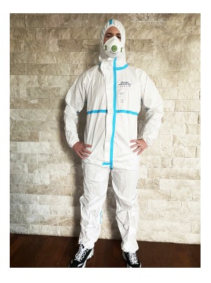 Osoba w białym kombinezonie ochronnym z niebieskimi akcentami i maską filtrującą na tle ściany z cegły.