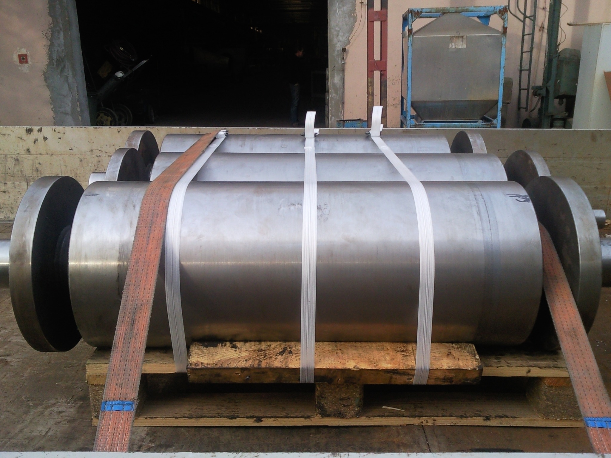 Szlifowane, stalowe walce zapakowane na drewnianej palecie, zabezpieczone taśmami transportowymi, w tle hala produkcyjna.