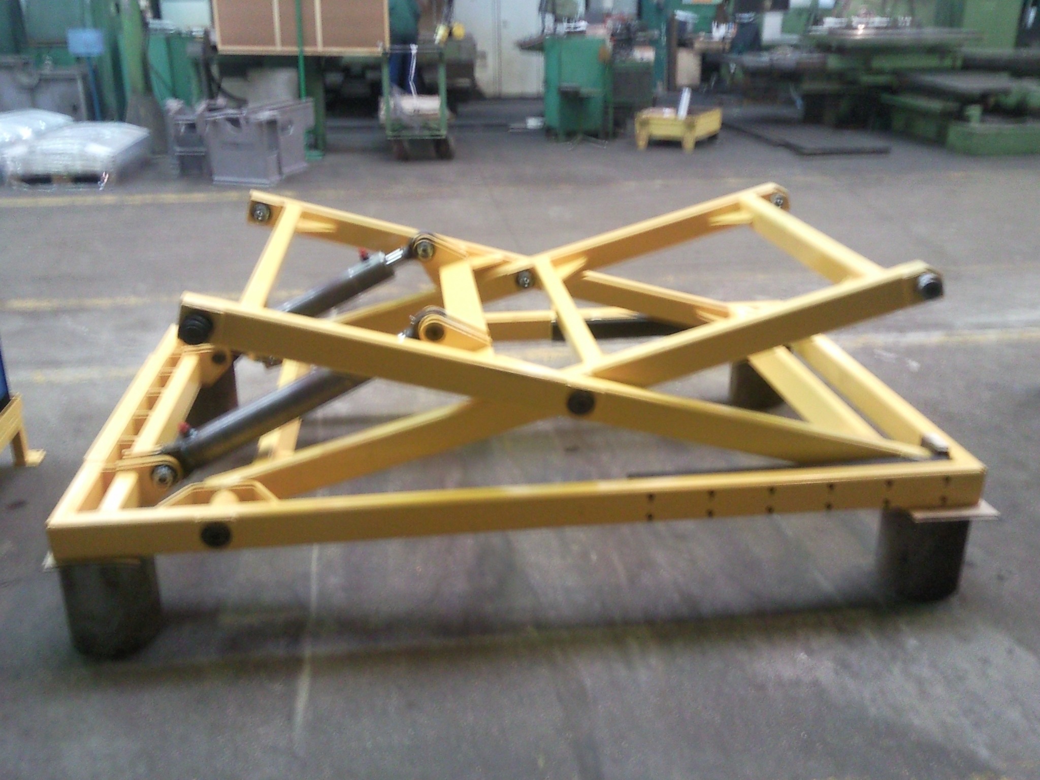 Żółta, stalowa konstrukcja nożycowa podnośnika hydraulicznego na betonowej posadzce hali przemysłowej, widoczne cylindry hydrauliczne i elementy mocujące.