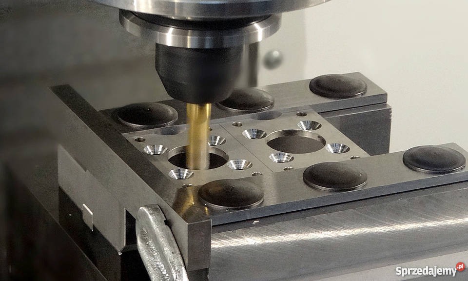 Precyzyjne wiercenie otworu w metalowej płycie za pomocą tokarki CNC, widoczne czarne podkładki stabilizujące i uchwyt mocujący.