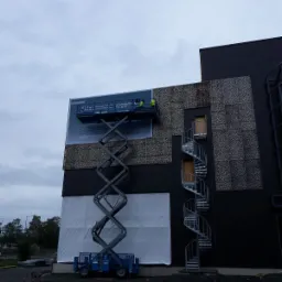 Montaż baneru reklamowego na elewacji budynku przez dwóch pracowników na podnośniku koszowym, widoczna spiralna klatka schodowa i fragment elewacji z panelami dekoracyjnymi.