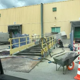 Pracownik w kombinezonie ochronnym i kasku, szlifuje metalową rampę w pobliżu magazynu z numerem 3, widoczna taczka z pomarańczowym kołem i stożek ostrzegawczy.