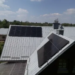 Panele słoneczne na metalowym dachu domu jednorodzinnego, widok z góry, błękitne niebo z chmurami w tle.