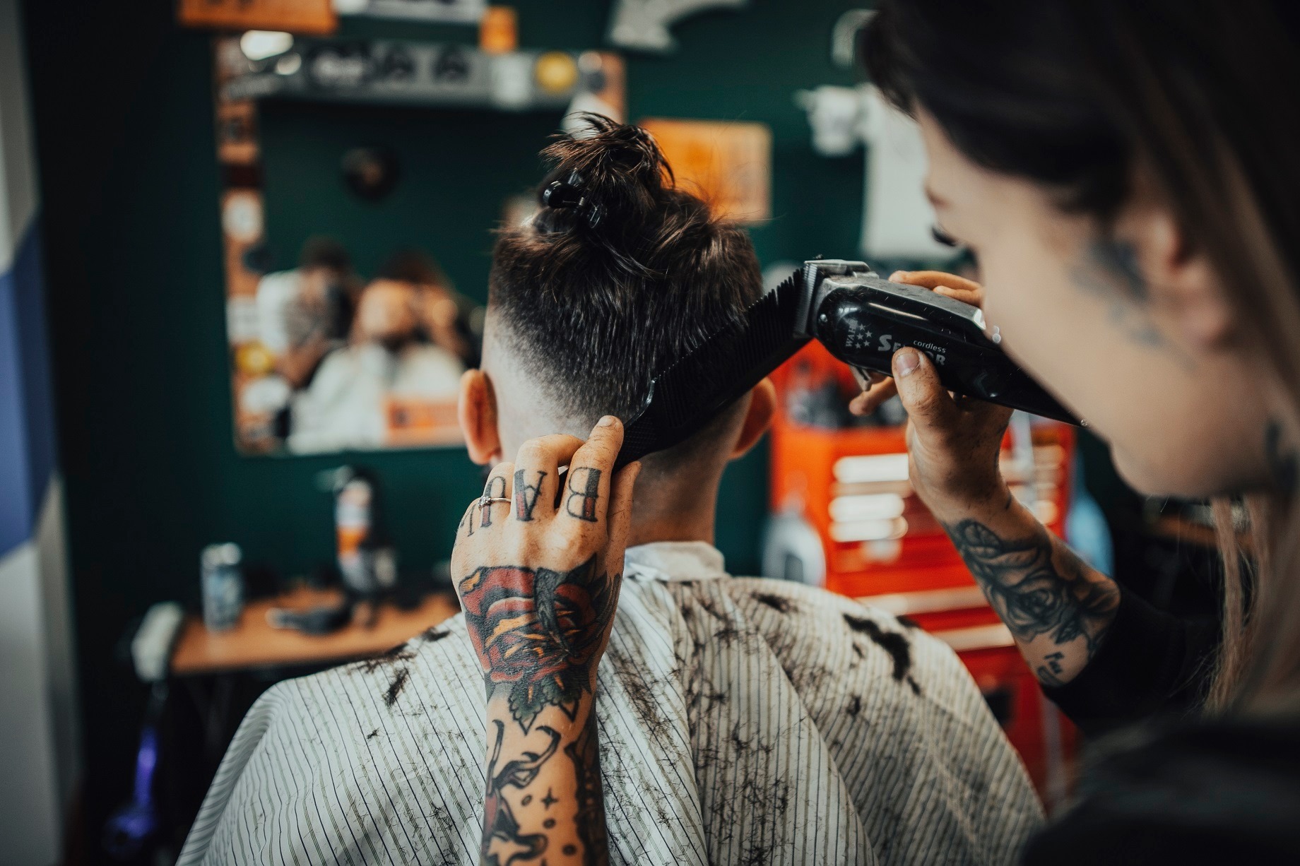 Strzyżenie męskie maszynką w salonie, widok z tyłu na klienta z włosami spiętymi w kitkę, tatuaże na dłoni i przedramieniu fryzjera.