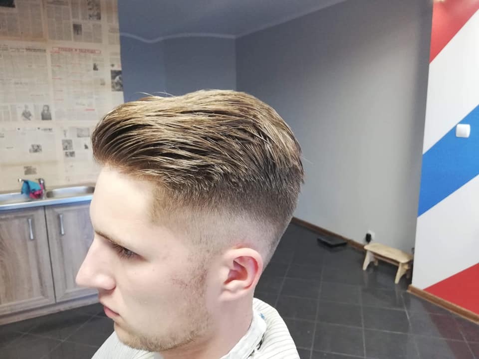 Męska fryzura z wycieniowanymi bokami, widoczna z profilu w salonie z gazetową tapetą i elementami barber shopu.