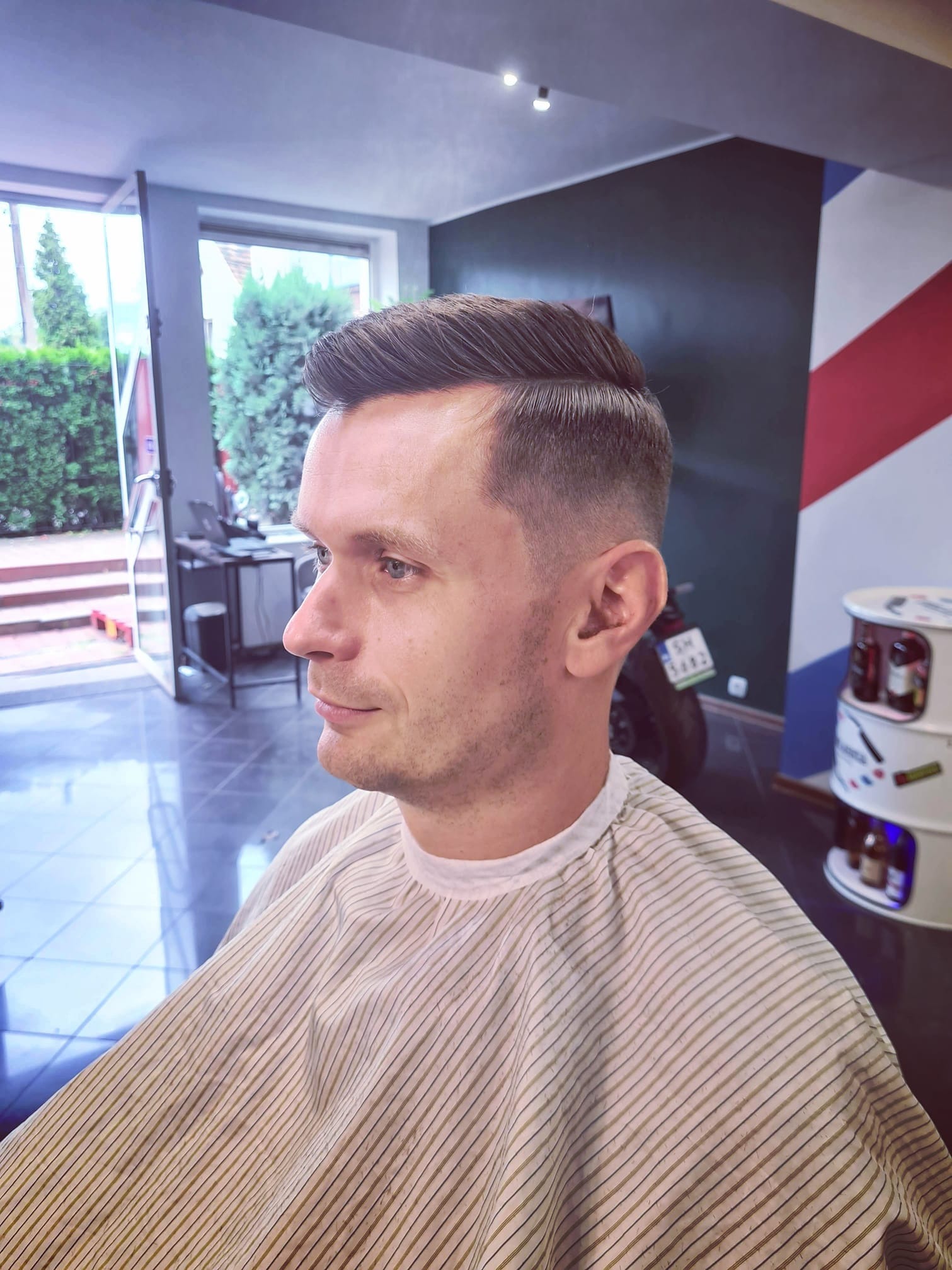 Mężczyzna z profilu w pelerynie fryzjerskiej po strzyżeniu z wygolonym bokiem i zaczesaną do tyłu górą, w tle widoczny motocykl i dekoracyjna ściana w salonie fryzjerskim.