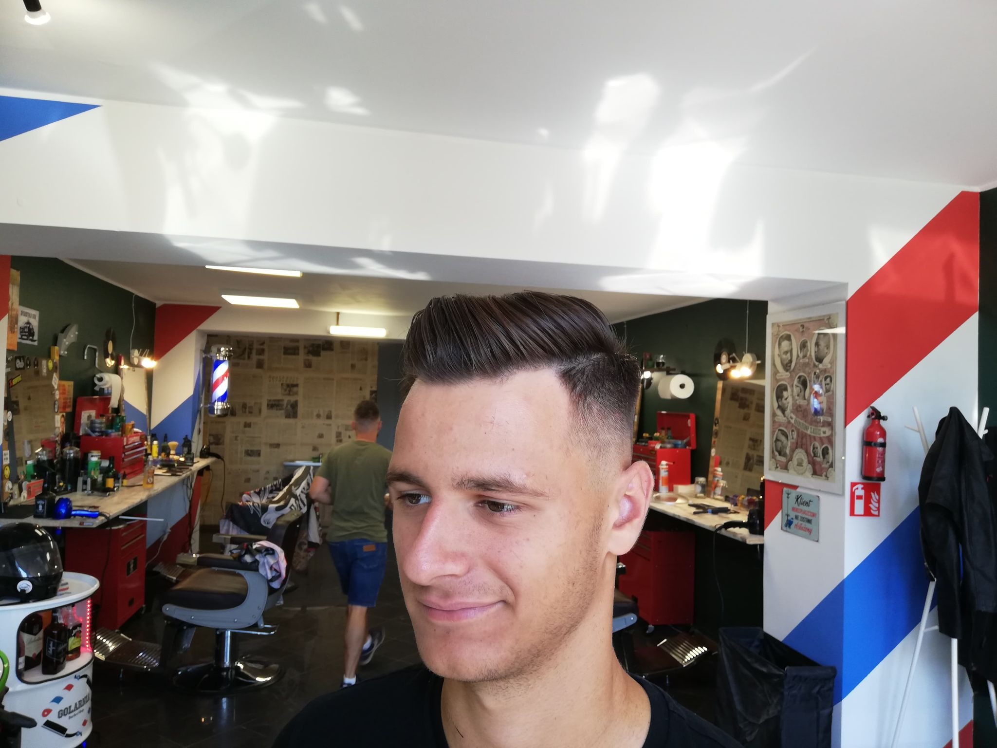 Młody mężczyzna z modną fryzurą typu fade w salonie fryzjerskim, widoczny w tle barber pole i stanowiska pracy.