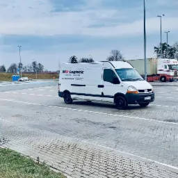 Biały bus z logo firmy logistycznej zaparkowany na wybrukowanym parkingu z widocznymi ciężarówkami w tle.