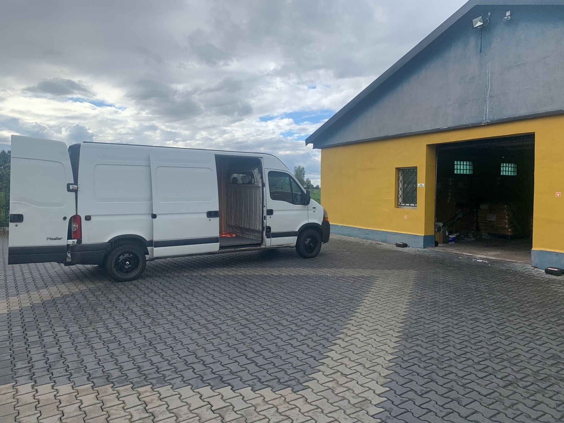 Biały bus z otwartymi drzwiami, zaparkowany na kostce brukowej przed żółtym budynkiem magazynowym w Elblągu, widoczny fragment wnętrza pojazdu.