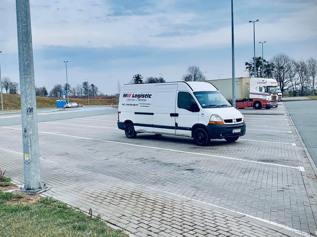 Biały bus z logo firmy logistycznej zaparkowany na wybrukowanym parkingu z widocznymi ciężarówkami w tle.