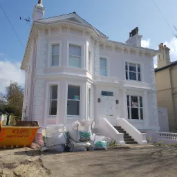 Odnowiony, dwupiętrowy dom w odcieniach różu i bieli, z widocznymi workami z materiałami budowlanymi i kontenerem na gruz przed budynkiem, sugerującymi trwający proces renowacji.