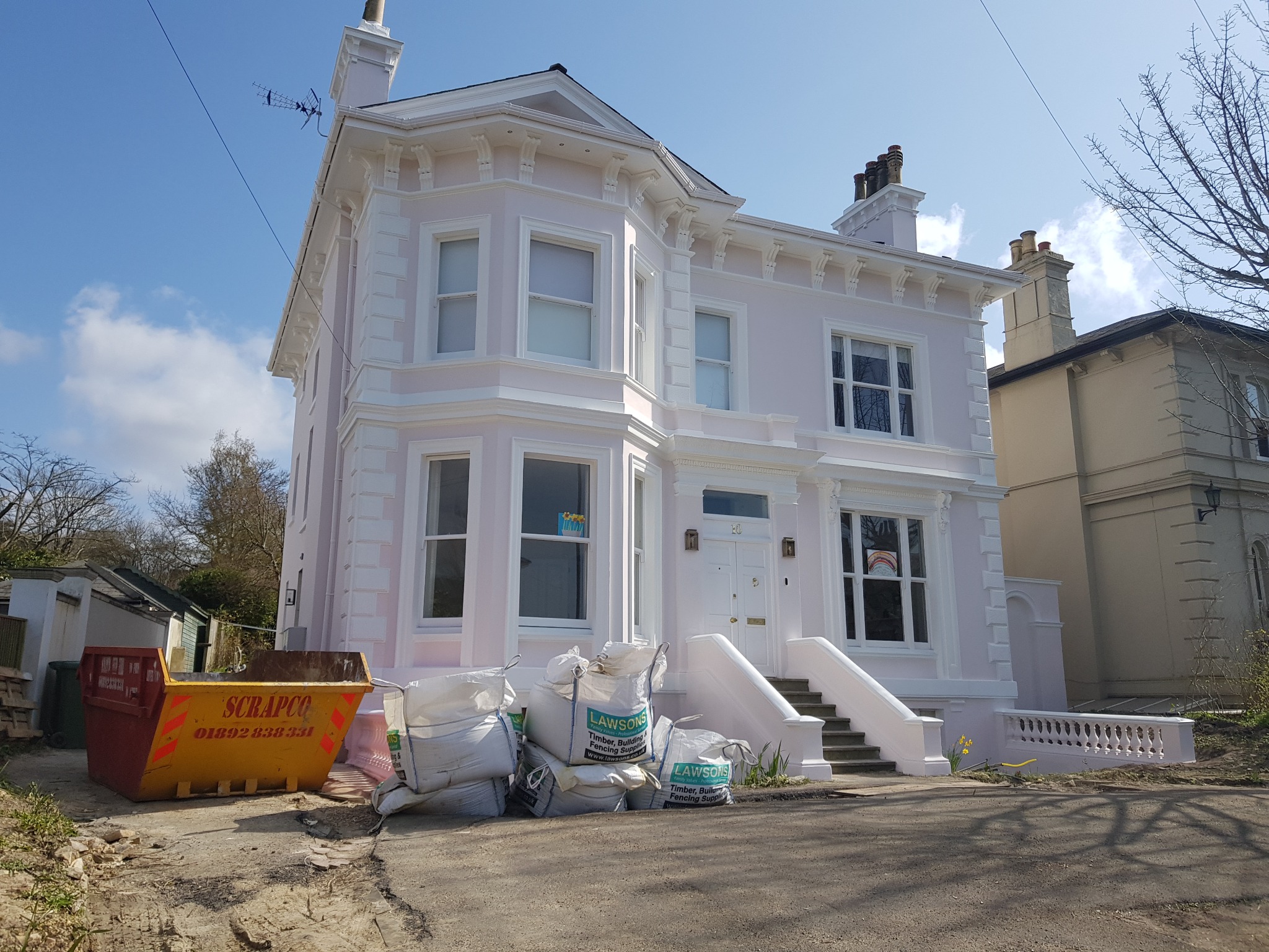 Odnowiony, dwupiętrowy dom w odcieniach różu i bieli, z widocznymi workami z materiałami budowlanymi i kontenerem na gruz przed budynkiem, sugerującymi trwający proces renowacji.