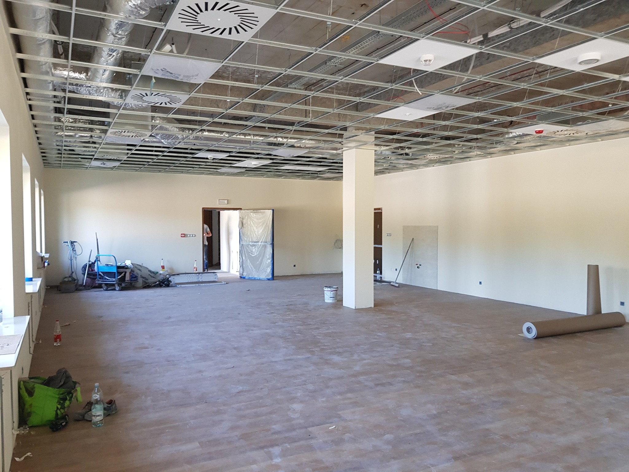 Przestronne wnętrze w trakcie prac wykończeniowych, widoczna metalowa konstrukcja sufitu podwieszanego, podłoga z paneli imitujących drewno, ściany w jasnym odcieniu, narzędzia i materiały...