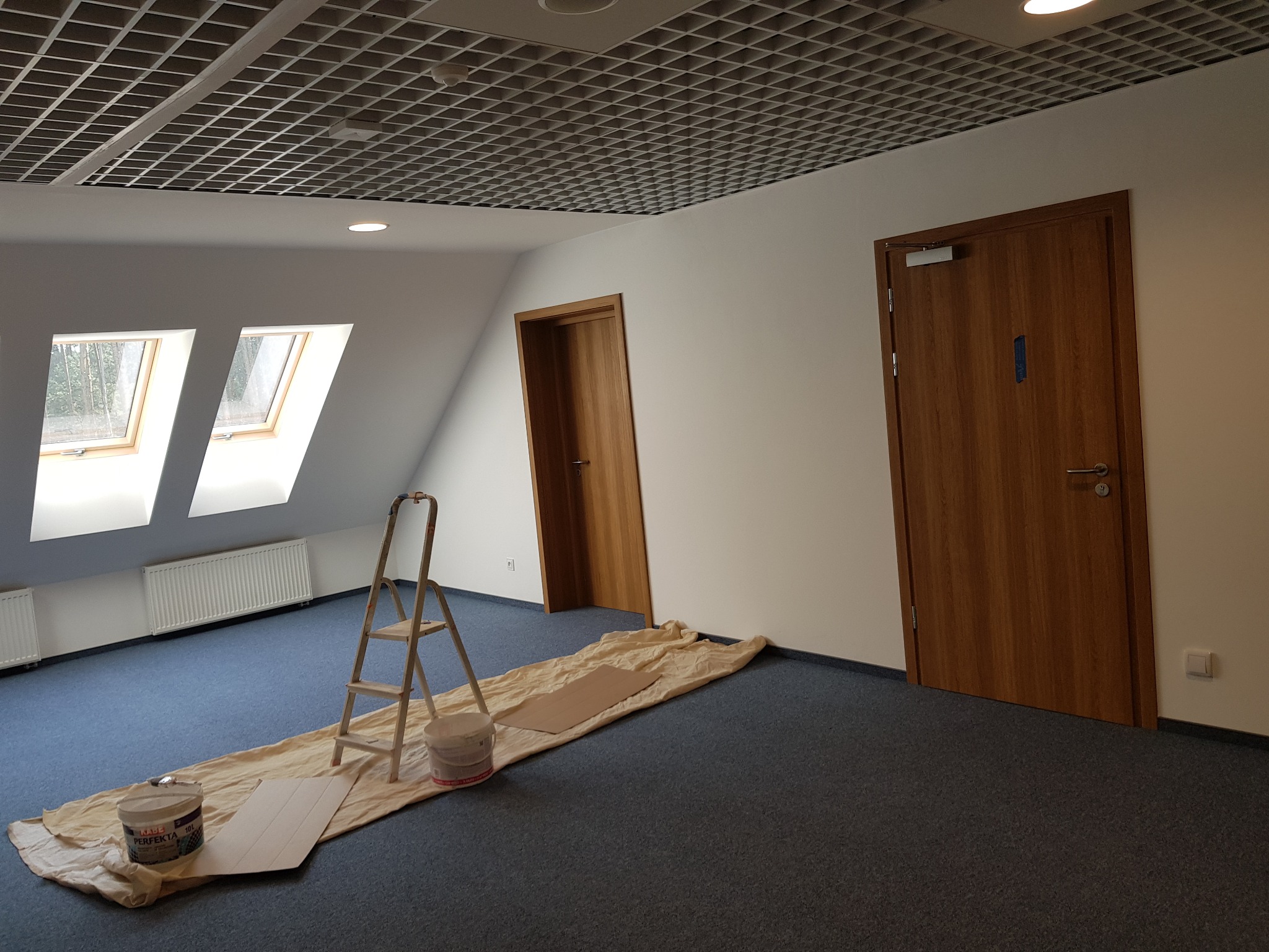 Przygotowania do malowania biura: rozstawiona drabina, wiadro z farbą KABE PERFEKTA, folia ochronna na granatowej wykładzinie, widoczne okna dachowe i krata wentylacyjna na suficie.