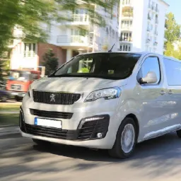 Srebrny bus Peugeot Expert w ruchu na tle drzew i budynku mieszkalnego, z widocznym w oddali wozem strażackim.