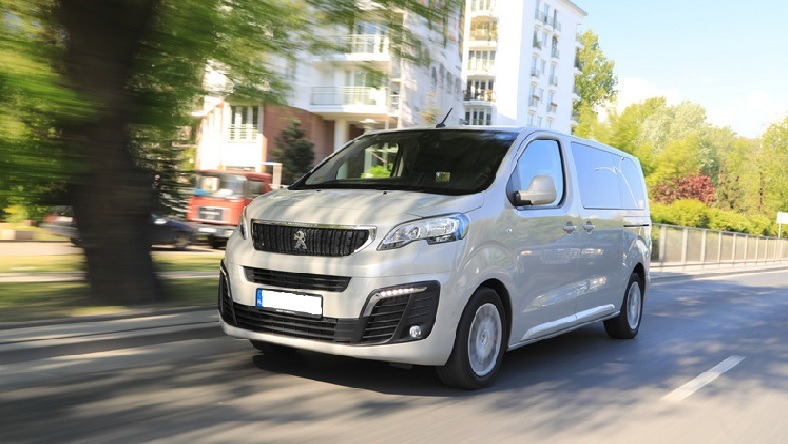 Srebrny bus Peugeot Expert w ruchu na tle drzew i budynku mieszkalnego, z widocznym w oddali wozem strażackim.