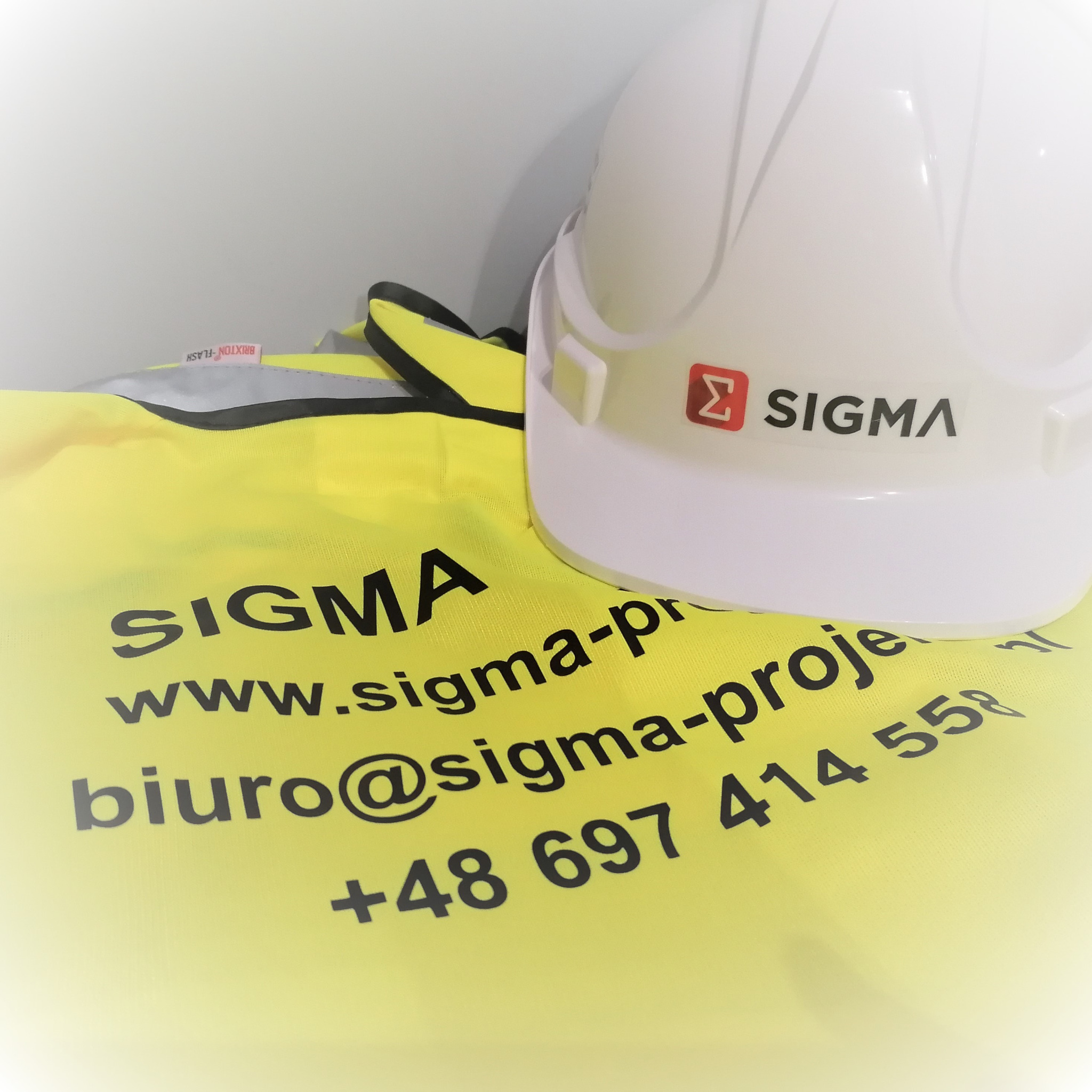 Żółta kamizelka odblaskowa z nadrukowanym logo, adresem strony internetowej, adresem email i numerem telefonu firmy Sigma Project, obok biały kask ochronny z logo firmy.