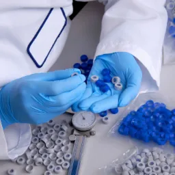 Pracownik w białym fartuchu i niebieskich rękawiczkach sortuje i mierzy suwmiarką plastikowe nakrętki z niebieskimi i szarymi elementami, obok woreczki z dużą ilością tych samych nakrętek.