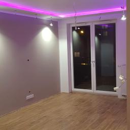 ewe - Pokój po remoncie z drewnopodobną podłogą, szarą ścianą i podwieszanym sufitem z fioletowym oświetleniem LED, widoczne niedokończone podłączenia lamp