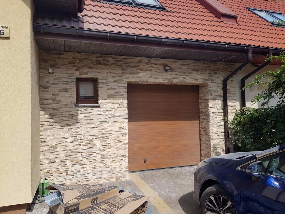 Elewacja budynku z jasnego kamienia dekoracyjnego, brązowa brama garażowa, małe okno z brązową ramą, widoczne kartony z opakowaniem kamienia dekoracyjnego, fragment samochodu.
