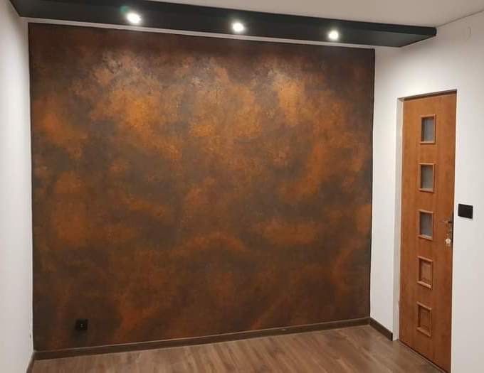 Ściana z efektem rdzy i drewniana podłoga w nowoczesnym wnętrzu, oświetlenie punktowe w czarnym suficie podwieszanym, brązowe drzwi z przeszkleniami.