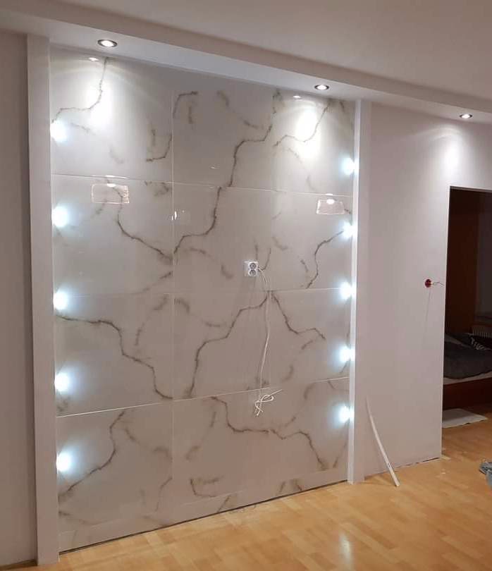 Ściana w łazience wyłożona białymi płytkami imitującymi marmur, z wbudowanym oświetleniem punktowym po bokach i górze, widoczne gniazdko elektryczne i przewody.