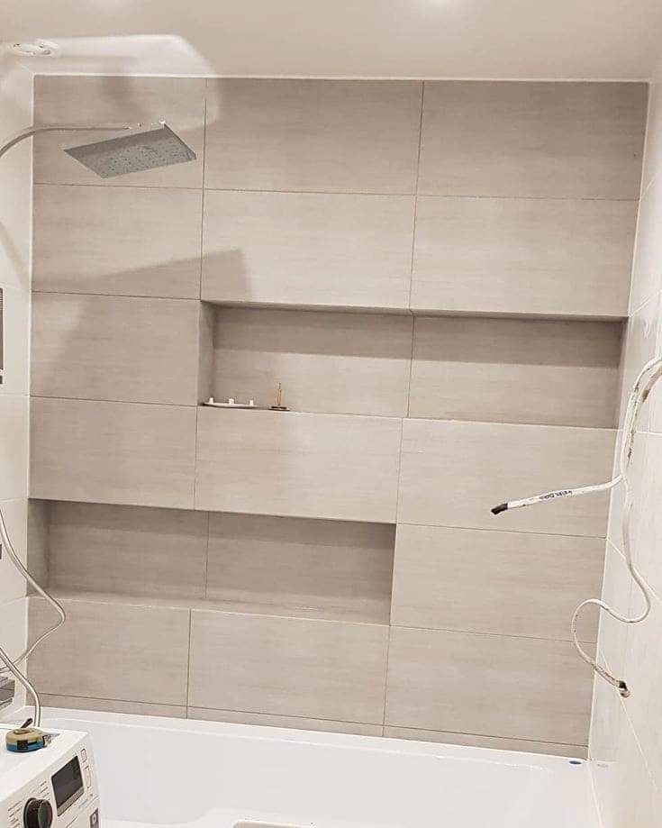 Beżowe, prostokątne płytki w łazience, ułożone z wnękami na kosmetyki, widoczny prysznic typu deszczownica i fragment wanny, kable wystające ze ściany wskazują na prace w toku.