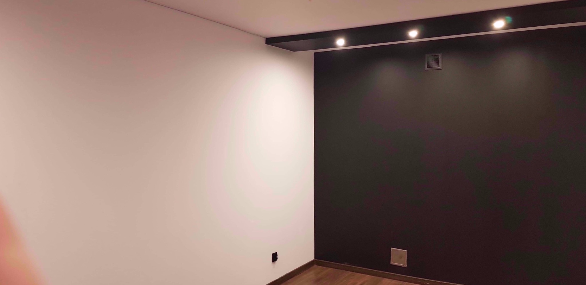 Pomieszczenie po remoncie z białą ścianą po lewej i ciemną, prawie czarną ścianą po prawej, z zamontowanym oświetleniem punktowym na suficie i drewnopodobną podłogą.