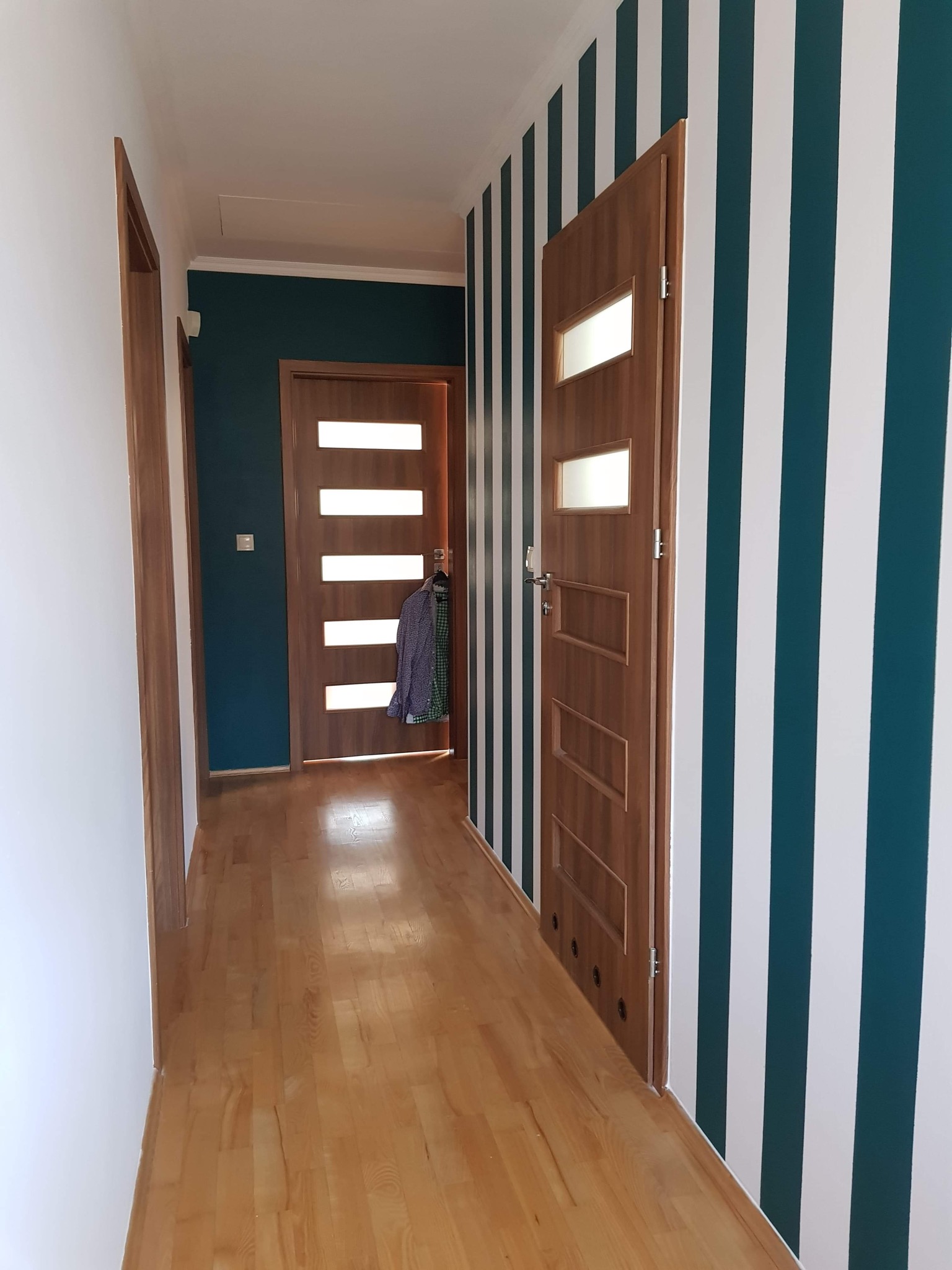 Wnętrze korytarza z drewnianą podłogą, drzwiami z przeszkleniami i ścianami w pionowe, biało-zielone pasy. Widoczna wisząca koszula na drzwiach w tle.