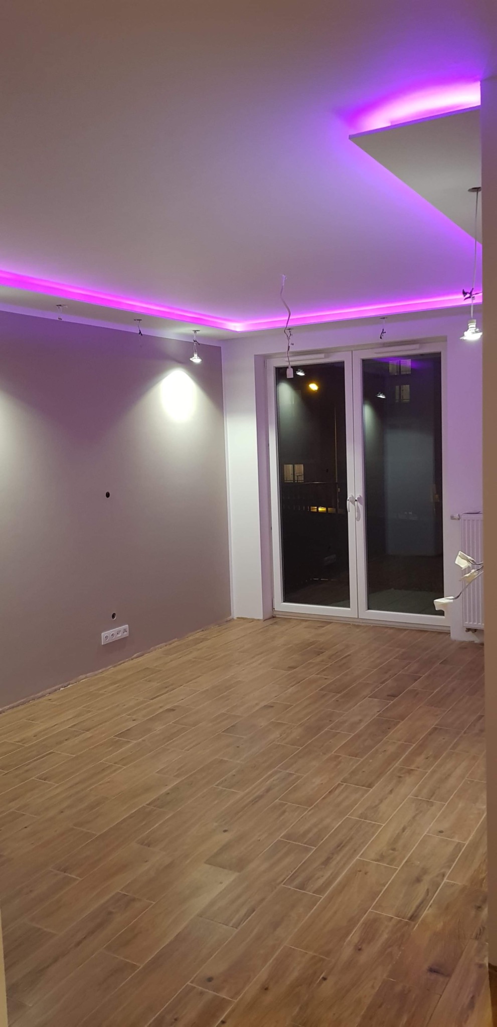 Pokój po remoncie z drewnopodobną podłogą, szarą ścianą i podwieszanym sufitem z fioletowym oświetleniem LED, widoczne niedokończone podłączenia lamp
