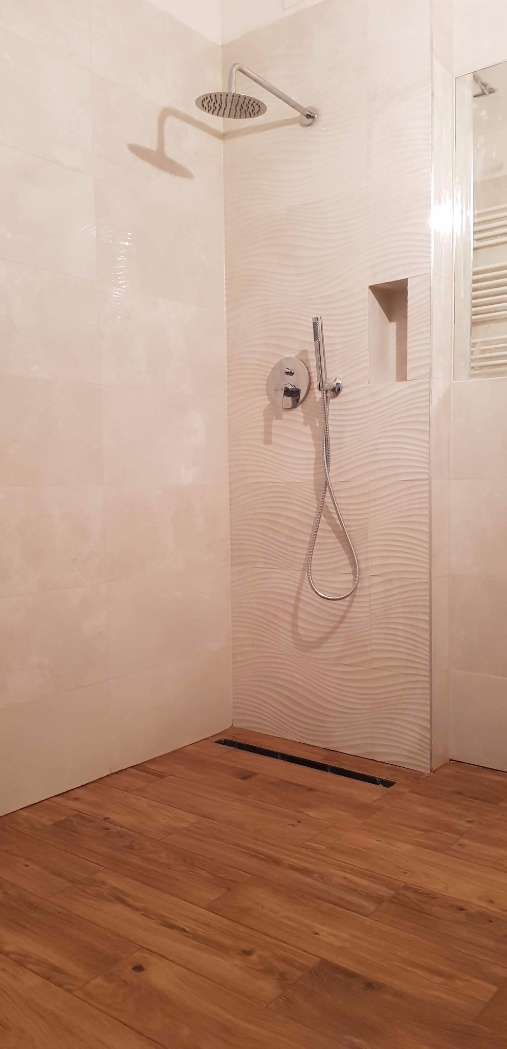 Nowoczesna kabina prysznicowa z drewnopodobną podłogą, deszczownicą i odpływem liniowym w posadzce, wykończona beżowymi płytkami z delikatnym wzorem fal.