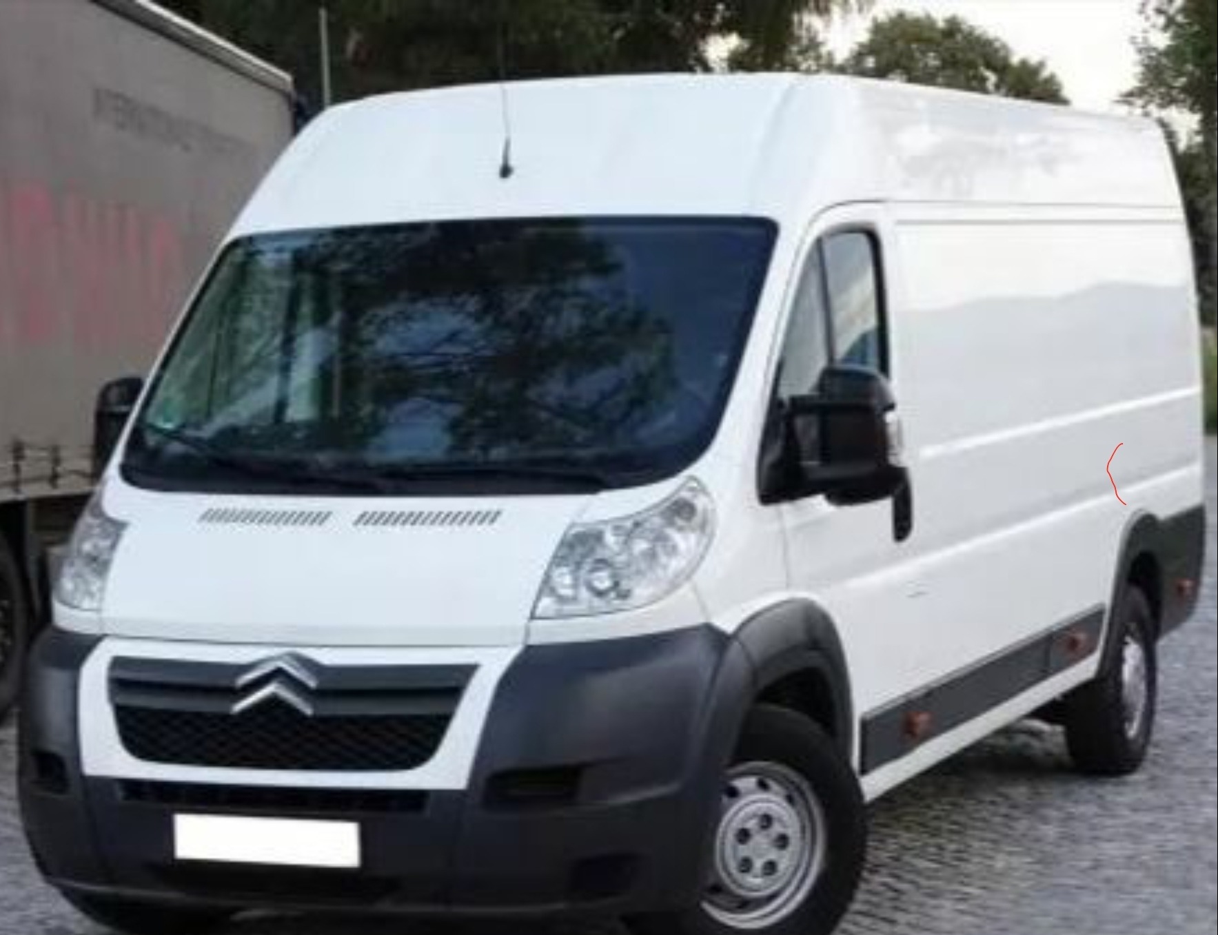 Biały samochód dostawczy Citroen z wysokim dachem, zaparkowany na zewnątrz, widok z przodu i z boku, z widocznym fragmentem naczepy innego pojazdu w tle.