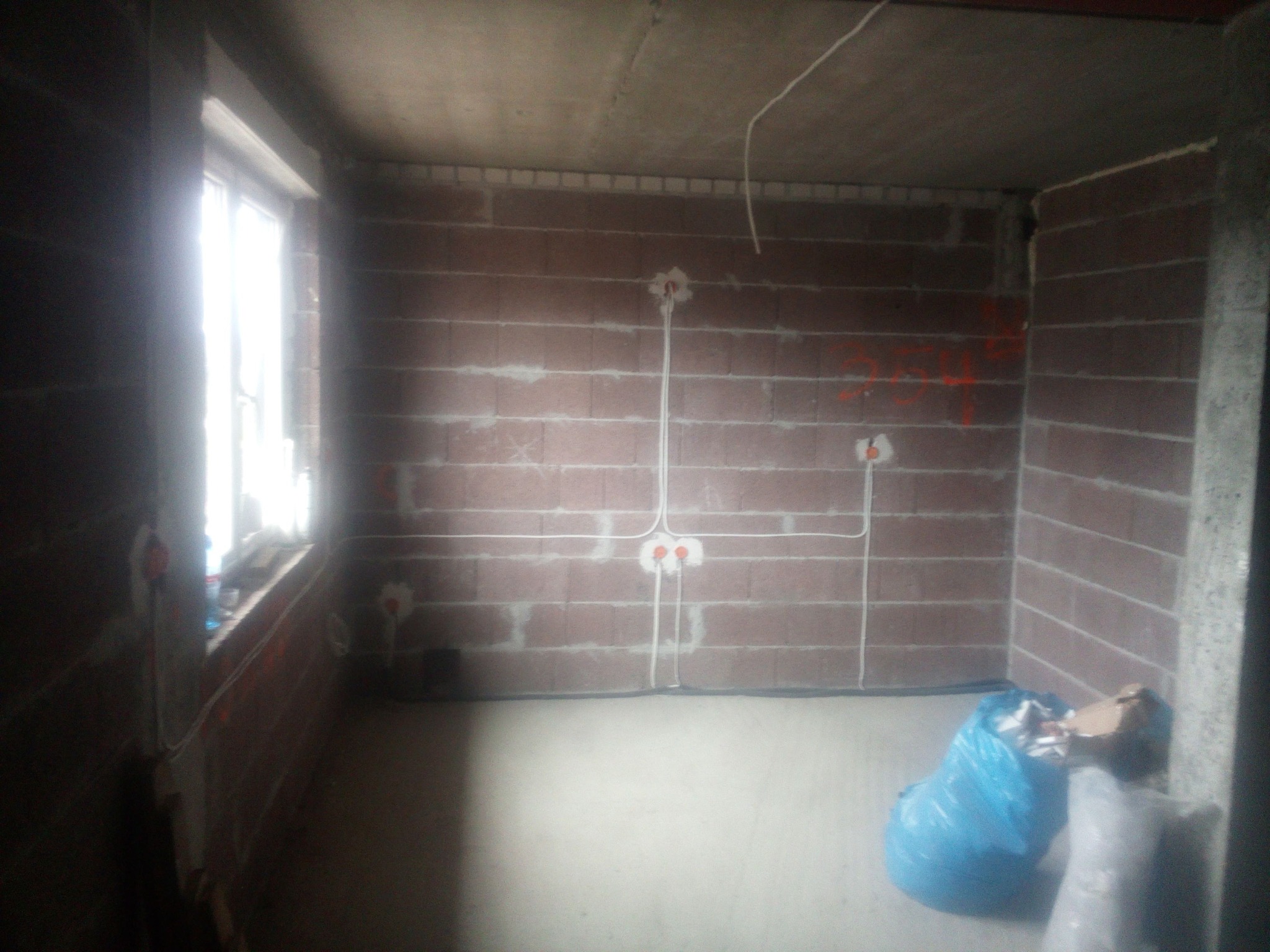 Surowy stan wnętrza z widoczną instalacją elektryczną w bruzdach na ceglanej ścianie oraz wystającymi przewodami z puszek, w rogu leżą worki na śmieci.