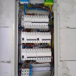 Rozdzielnica elektryczna z zainstalowanymi bezpiecznikami automatycznymi i okablowaniem, zamontowana w surowej ścianie z betonu komórkowego.