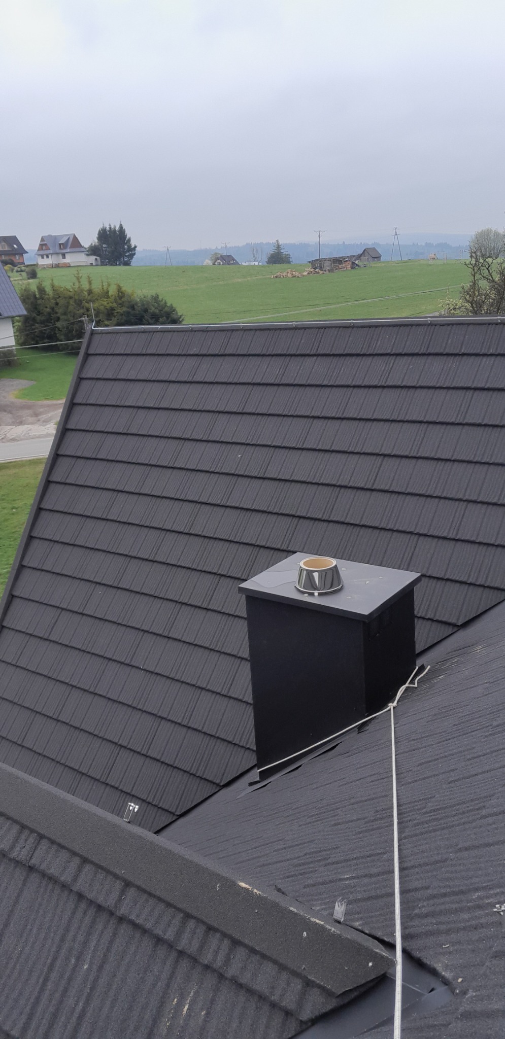 Czarny komin z metalową nasadą na dachu z ciemnej blachy dachówkowej, widok z góry na okoliczne domy i pola w pochmurny dzień.