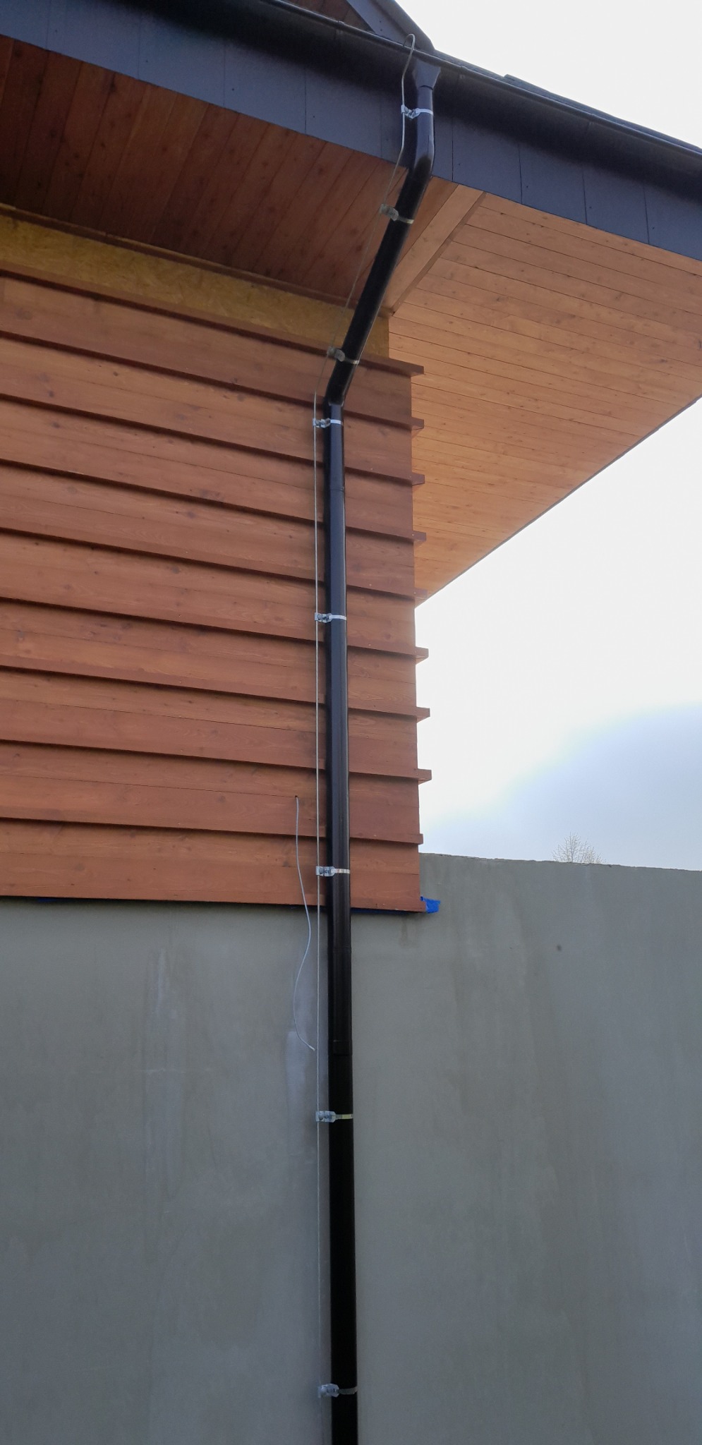 Czarna rura spustowa przymocowana do brązowej drewnianej elewacji domu za pomocą metalowych uchwytów, z widocznym białym kablem biegnącym wzdłuż rury.