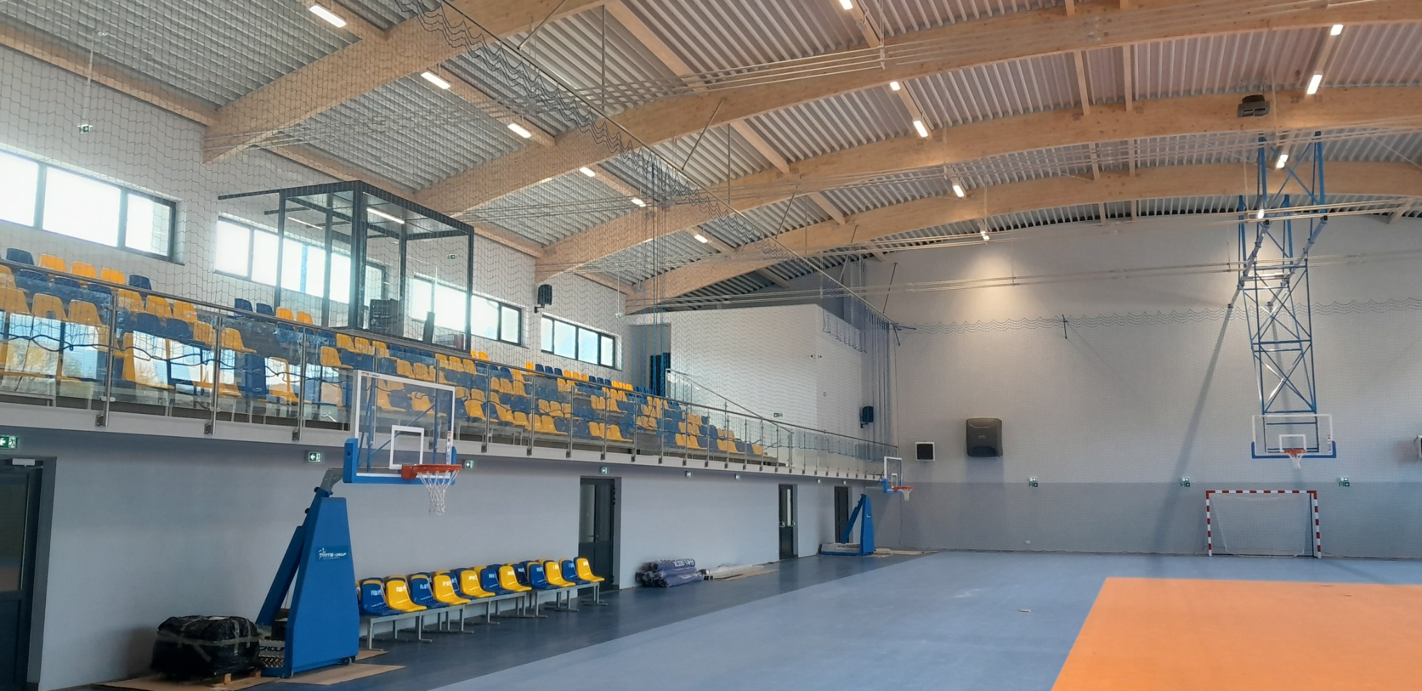 Wnętrze nowej hali sportowej z trybunami w kolorach żółto-niebieskich, drewnianą konstrukcją dachu i zainstalowanym oświetleniem, widoczne kosze do koszykówki i bramka do piłki nożnej.